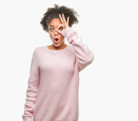 Fototapeta na wymiar Young afro american woman wearing winter sweater over isolated background doing ok gesture shocked with surprised face, eye looking through fingers. Unbelieving expression.