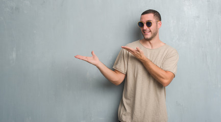 Young caucasian man over grey grunge wall wearing sunglasses amazed and smiling to the camera while presenting with hand and pointing with finger.