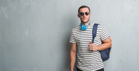 Young caucasian student man over grey grunge wall wearing headphones and backpack with a confident expression on smart face thinking serious