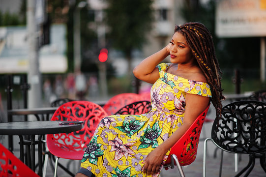 Cute small height african american girl with dreadlocks, wear at coloured yellow dress, sitting at outdoor cafe on red chair.