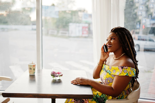 Cute small height african american girl with dreadlocks, wear at coloured yellow dress, sitting at cafe and speaking on phone.