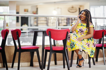 Cute small height african american girl with dreadlocks, wear at coloured yellow dress, sitting on cafe at shopping center and speaking on phone.