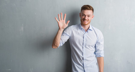 Young redhead business man over grey grunge wall showing and pointing up with fingers number five while smiling confident and happy.