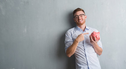 Young redhead man over grey grunge wall holding piggy bank very happy pointing with hand and finger