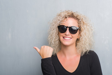 Young blonde woman with curly hair over grunge grey background smiling with happy face looking and pointing to the side with thumb up.