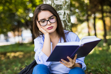 Young woman in eyeglasses sitting on grass under tree and reading book in park