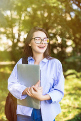 Fototapeta premium Smiling brunette student wearing glasses and shirt walking outdoor with laptop and backpack background of green park