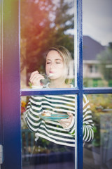 Young girl with pleasure eating a cake near a window at home.