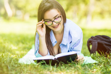 Fototapeta na wymiar Young nice attentive woman in glasses lies on green grass and reads book against city park.
