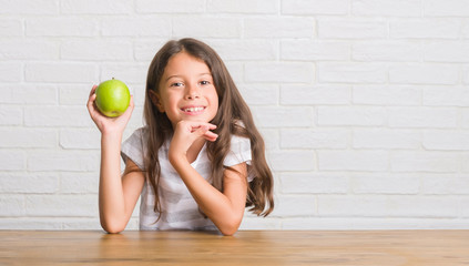 Young hispanic kid sitting on the table eating fresh green apple with a happy face standing and smiling with a confident smile showing teeth