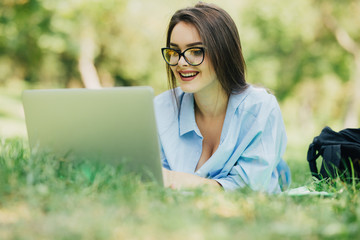 Young woman in eyesglasses lying on grass in park and using laptop