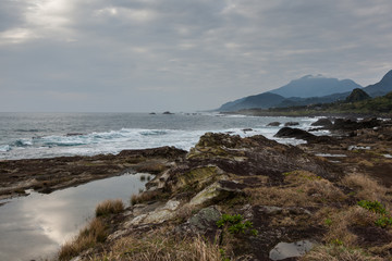 Fototapeta na wymiar Taiwan East Coast Rocky Coastline Background Image - Overcast Skies, Exotic Rock Formations, Grass and Waves in the Ocean. Ocean Coastline, Asia Landscape Photography, Rays of sunshine in background