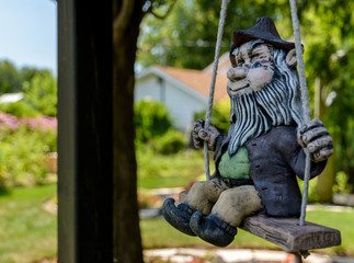 Rustic Looking Male Gnome With A White Beard And A Hat On A Swing With Gardens In The Background
