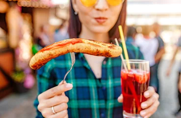 Tuinposter Young happy asian traveler woman eating fried sausage and drinks mug of beer at the fair market square in Germany, beer and food festival concept © EdNurg