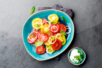 Red and yellow fresh tomato salad with onion, basil and white sauce on blue plate. Gray slate background, top view