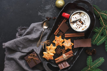 A mug of hot chocolate with marshmallows, star cookies, spices, chocolate slices, fir branches on a black concrete background. Christmas background. View from above, flat lay.