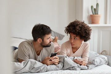 Lovely mixed race couple have pleasant talk, use mobile phones in bed at morning, look seriously at each other, enjoy coziness at bedroom, free wifi connection at home. Contemporary technology