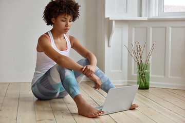 Horizontal shot of sporty dark skinned fitness girl dressed in casual t shirt and leggings, watches tutorial video on laptop computer, sits on floor in empty spacious room, uses modern gadget