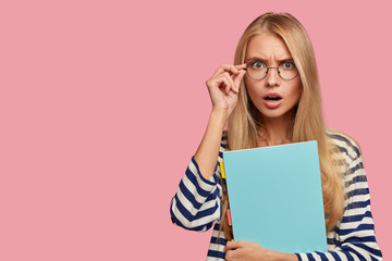 Portrait of terrified female keeps hand on rim of spectacles, dressed in sailor casual sweater,...
