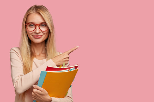Studio shot of good looking Caucasian teacher attracts your attention to copy space, holds books, wears round spectacles, explains material to pupils, points with index finger on pink background