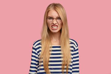 Portrait of irritated blonde woman clenches teeth angrily, expresses madness and fury, doesnt like something, has displeased annoyed facial expression, wears round glasses, isolated on pink. - obrazy, fototapety, plakaty