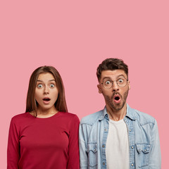 Vertical shot of stupefied young girl and boy stare with bugged eyes, have surprised face expressions, open mouthes from great amazement, isolated over pink background with copy space above.