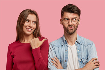 Photo of cheerful young woman points with thumb at hesitant boyfriend, suggests asks him about what happened, stand together against pink studio wall. Look at my displeased uncertain best friend