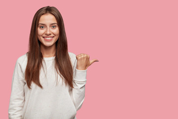 Cheerful positive young European woman with dark hair, broad shining smile, points with thumb...