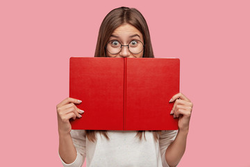 Happy book reader hides behind textbook, has joyful expression, finds out something new, has...