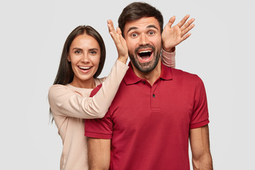 Overjoyed funny girlfriend and boyfriend foolish together, have happy expressions, pray fools indoor. Positive European woman makes ears to guy with both palms, have fun during weekend at home