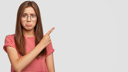 Confused troubled offensed woman points in displeasure at free space, dislikes something, wears...