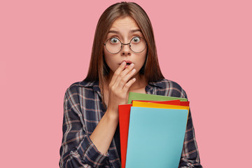 Isolated shot of scared young European woman geek keeps hands on opened mouth, has bugged eyes,...