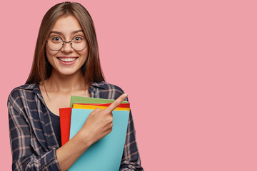Studio shot of good looking young woman with dark hair, dressed in checkered shirt, points with...