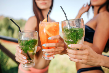 Fototapeta premium Cut view of three models stands and hold three glasses with cocktails. There are one orange and two green colored drinks. Young women keep them together.