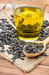 Glass cup of cooking oil and wooden spoon with sunflower seeds on napkin of burlap.