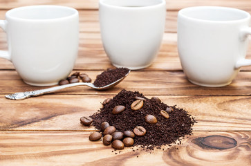 Heap of ground coffee and coffee beans with cups and spoon on wooden table.