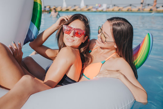 Two young models chilling on white float in swimming pool. First wear red suglasses. She holds hand on hair and keep eyes closed. Second one look at her and smile. Se also wears sunglasses. They have