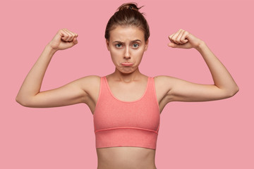Self detrermined fit woman with sporty body, shows muscles, wears casual top, purses lips, ready for workout and physical training, isolated over pink background. People and motivation concept - obrazy, fototapety, plakaty
