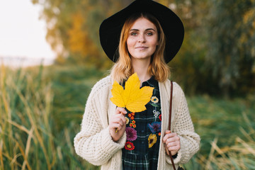 Hippie girl with a yellow maple leaf in a knitted sweater and hat walks autumn park.