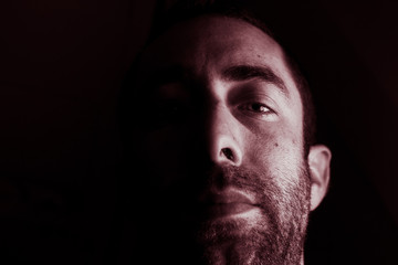 Dramatic portrait of a young man half expressionless face in deep shadow looking at camera isolated on black background