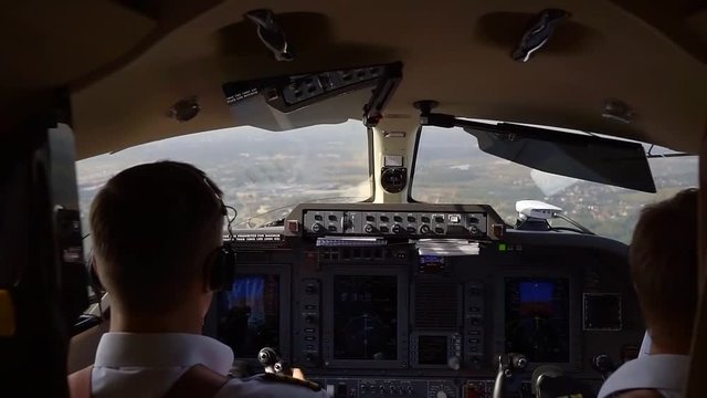 View from behind of two pilots in cockpit flying a plane, control panel with buttons and switches.