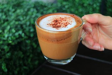 Closed Up a Cup of Hot Cappuccino Coffee Holding in Woman's Hand, with Blurred Green Bush in Background