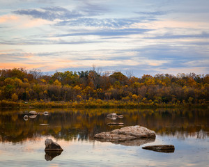 Sunset With A Forest With Fall Colours Along A River With Rocks And Reflections And Wispy Clouds