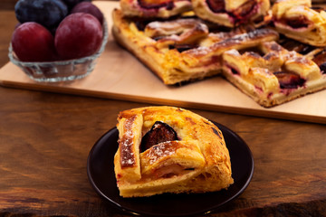 A piece of homemade puff pastry pie with plums on a round plate, wooden background, close-up.