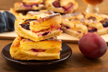 Cut pieces of puff pastry with cottage cheese, stacked on top of each other on a round plate, next to a wooden background ripe plum purple.