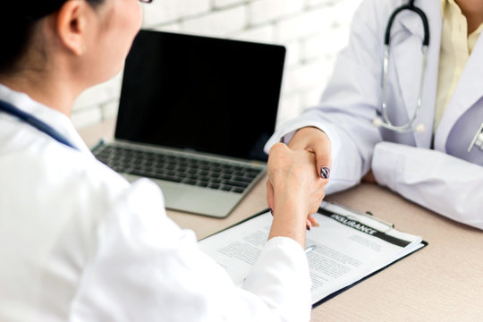 Close Up Of Two Doctors Woman Shaking Hands To Each Other Sitting At The Table In Hospital Office. Medicine And Trust. Concept Of Collaboration In Medicine.