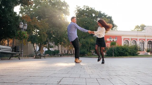 Young business couple on date, man hugs and spinning around woman. Girl jumps into boyfriend's arms, he whirling her at empty city street. Having fun together, fooling around.