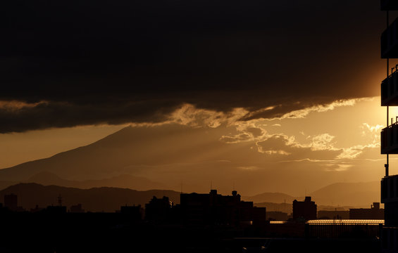 Sunlight flooding in below dark clouds during sunset over a small city with the outline of mount Fuji in the background
