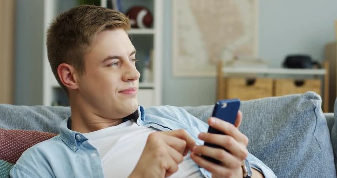 Young Caucasian man scrolling and taping on the smartphone and looking at the side in the living room.