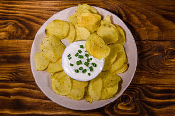 Ceramic plate with potato chips and glass bowl with sour cream on wooden table. Top view